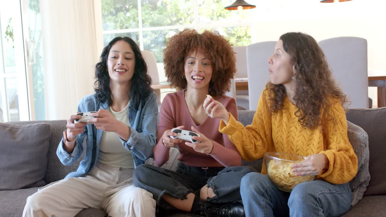 Unas amigas cogen los mandos mientras se carga el juego, jugando en un sofá gris, compartiendo patatas fritas y riendo.
