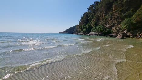 Primer-Plano-De-Las-Olas-Salpicando-En-La-Costa-Arenosa,-Cleef-Rocoso-Cubierto-De-Vegetación-Exuberante,-Playa-Paradisíaca,-Isla-De-Thassos,-Grecia