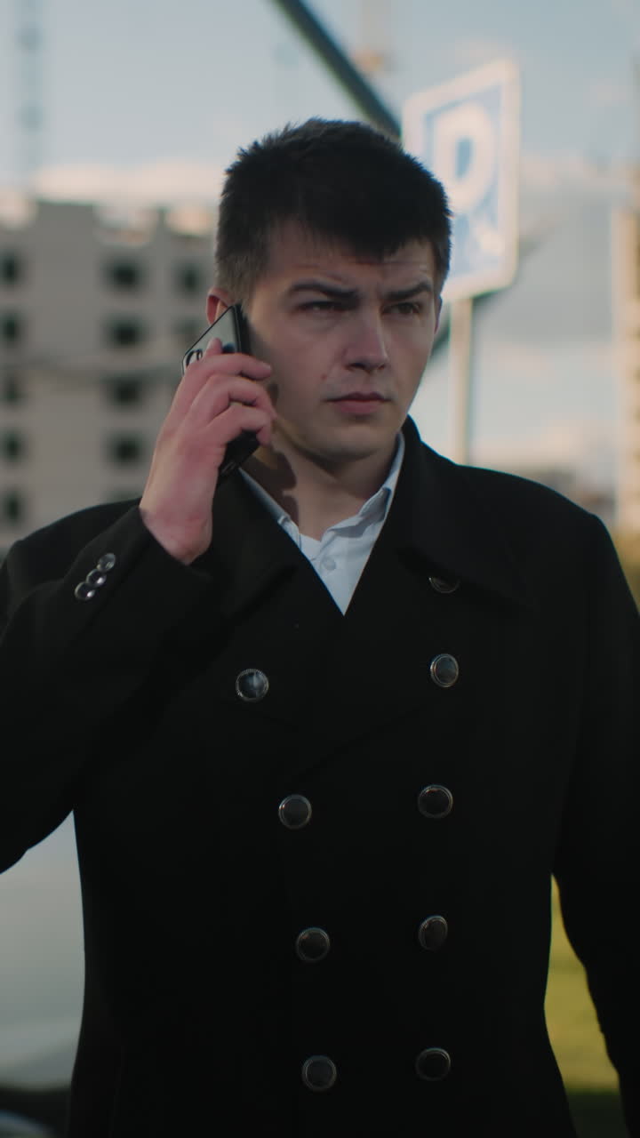 검은 코트를 입은 남자가 차를 떠나 심각한 표정으로 주머니에서 전화를 꺼내는 동안 주차된 자동차, 현대적인 건물, 움직이는 차량 및 초록색을 특징으로하는 활기찬 도시 지역에서