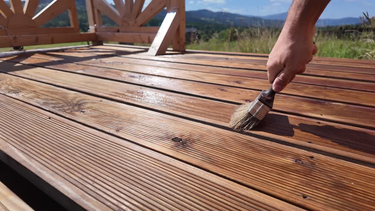 Working man oils varnishing impregnating wooden terrace boards. Decorative railing, serene blue sky, clouds, and mountain horizon evoke home improvement and outdoor maintenance themes - DIY