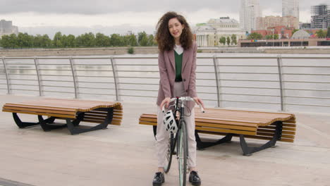 hermosa mujer rizada con ropa formal mirando y sonriendo a la cámara mientras se sienta en una bicicleta en el puente de la ciudad 1