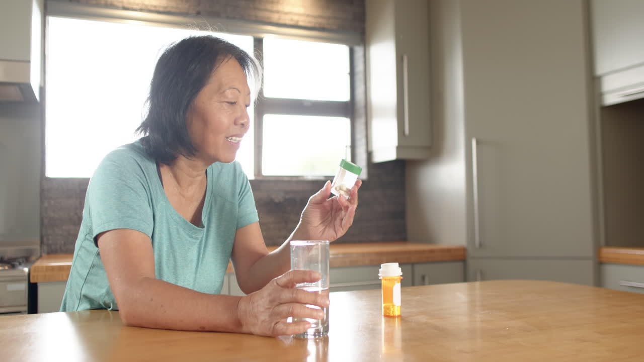 Senior Asian woman holding medication and glass of water at kitchen table, copy space