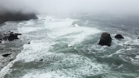 las olas blancas y espumosas de color verde esmeralda chocan en una costa rocosa y escarpada cubierta de niebla, aérea