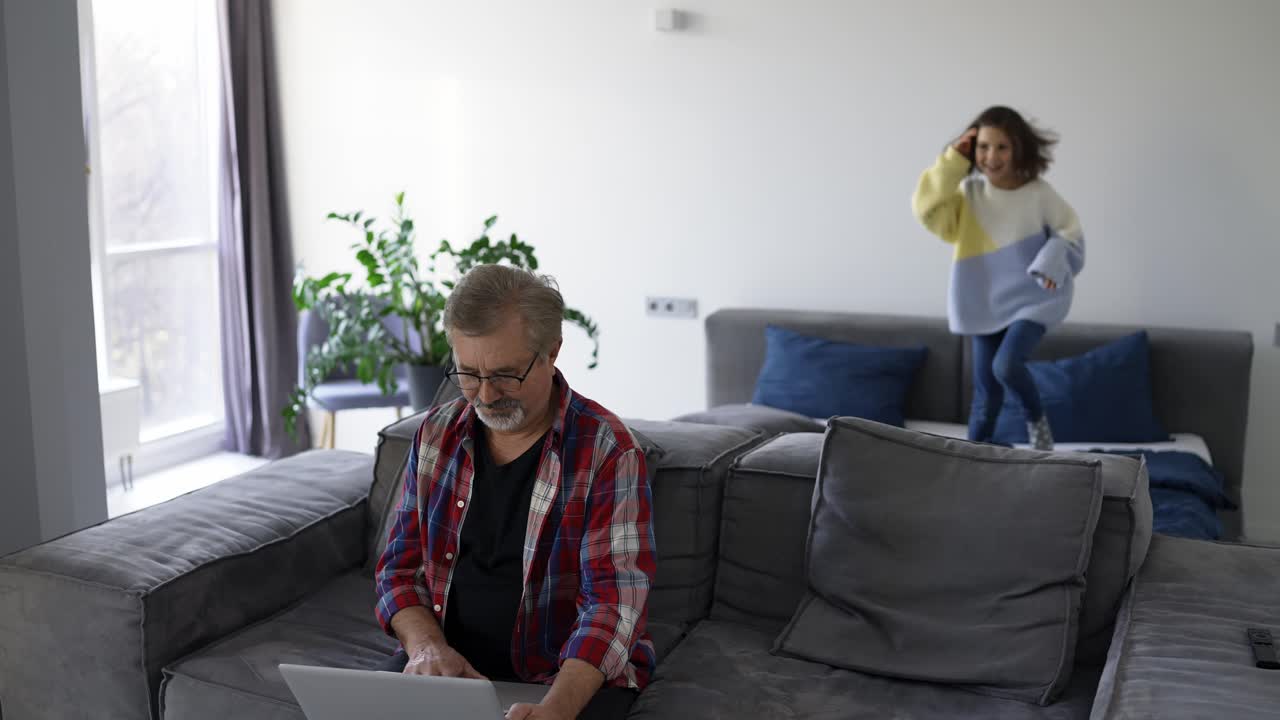 Adorable little girl playing on the background while grandfather using laptop at home