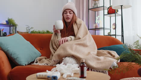 sick ill woman suffering from cold drinking hot tea sitting on couch sneezes wipes snot into napkin