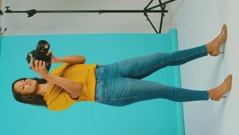 Vertical-Video-Of-Mature-Female-Photographer-With-Camera-Against-Blue-Background-On-Shoot-In-Studio