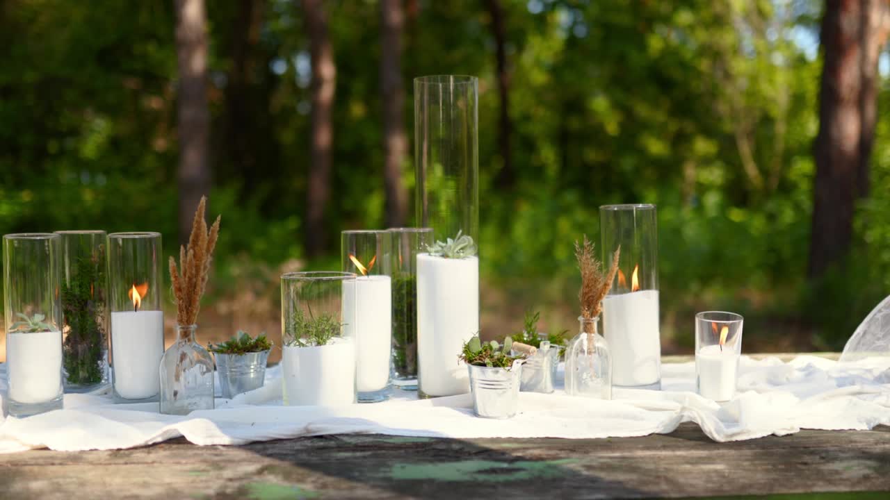 Wedding party banquet outdoors in pine forest. Dining tables decorated in boho style with candles in glass vases, white cloth, succulent flowers. Eco organic floral composition decor for holiday