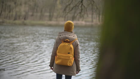 Ein-Junges-Mädchen-In-Einem-Wald,-Das-Eine-Gelbe-Wollmütze-Und-Einen-Gelben-Rucksack-Trägt-Und-Den-See-Betrachtet