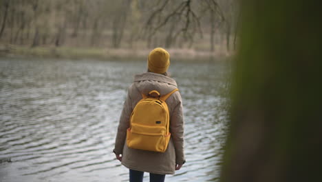 Una-Niña-En-Un-Bosque-Que-Usa-Un-Gorro-De-Lana-Amarillo-Y-Una-Mochila-Amarilla-Contemplando-El-Lago