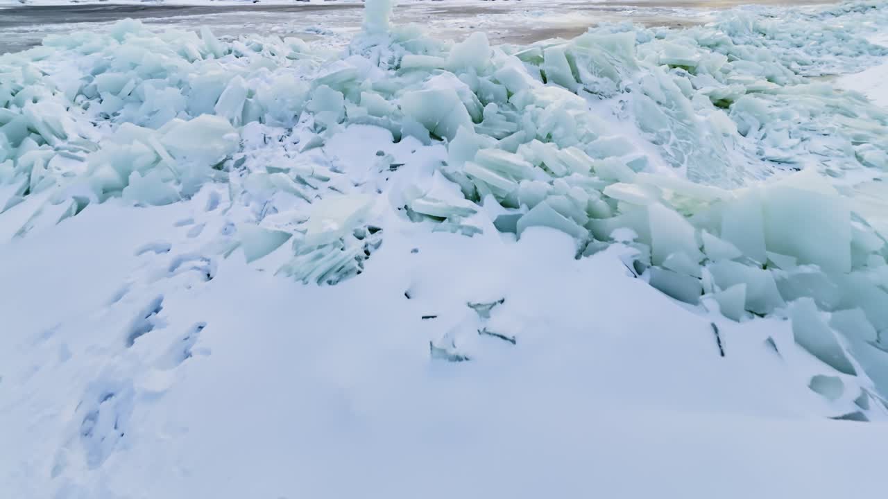 Low aerial shot over unstable winter ice with footprints crossing snow-covered slabs, dark melt channels, and piled broken ice in a hazardous frozen shoreline scene