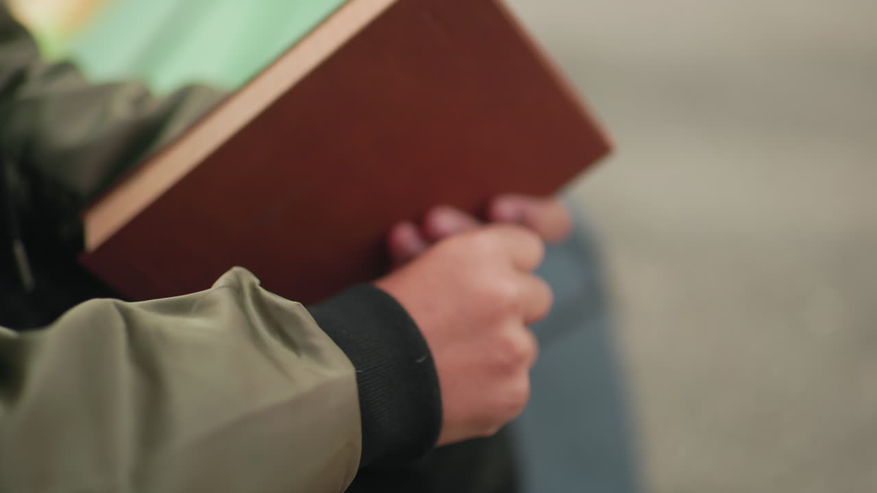 Close up partial view of someone seated holding brown cover book, gently hitting it on hand while resting outdoors with backpack nearby, wearing checkered pants and green jacket