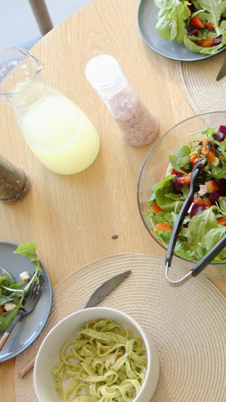 Vídeo vertical: Braço feminino com blusa rosa alcançando a mesa durante uma refeição, começando a levantar um copo e comendo salada.
