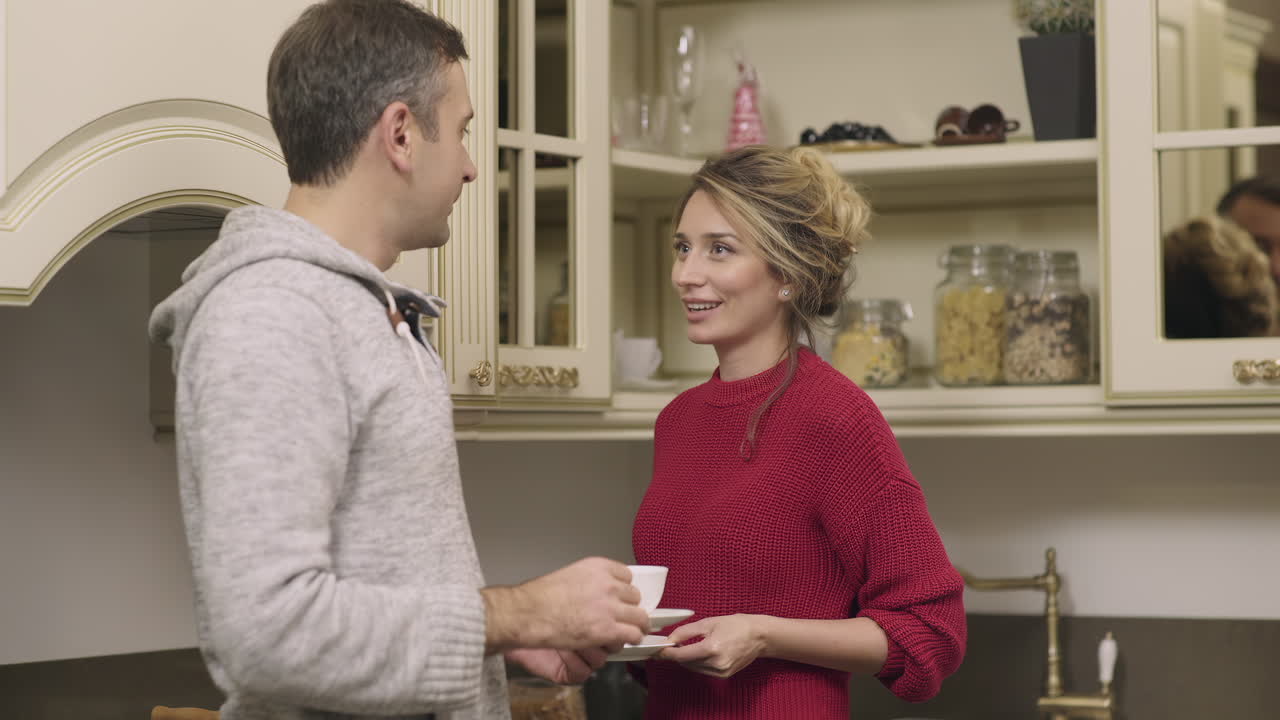 A man and woman share a warm interaction in a stylish kitchen, holding cups and smiling at each other while enjoying a relaxing afternoon