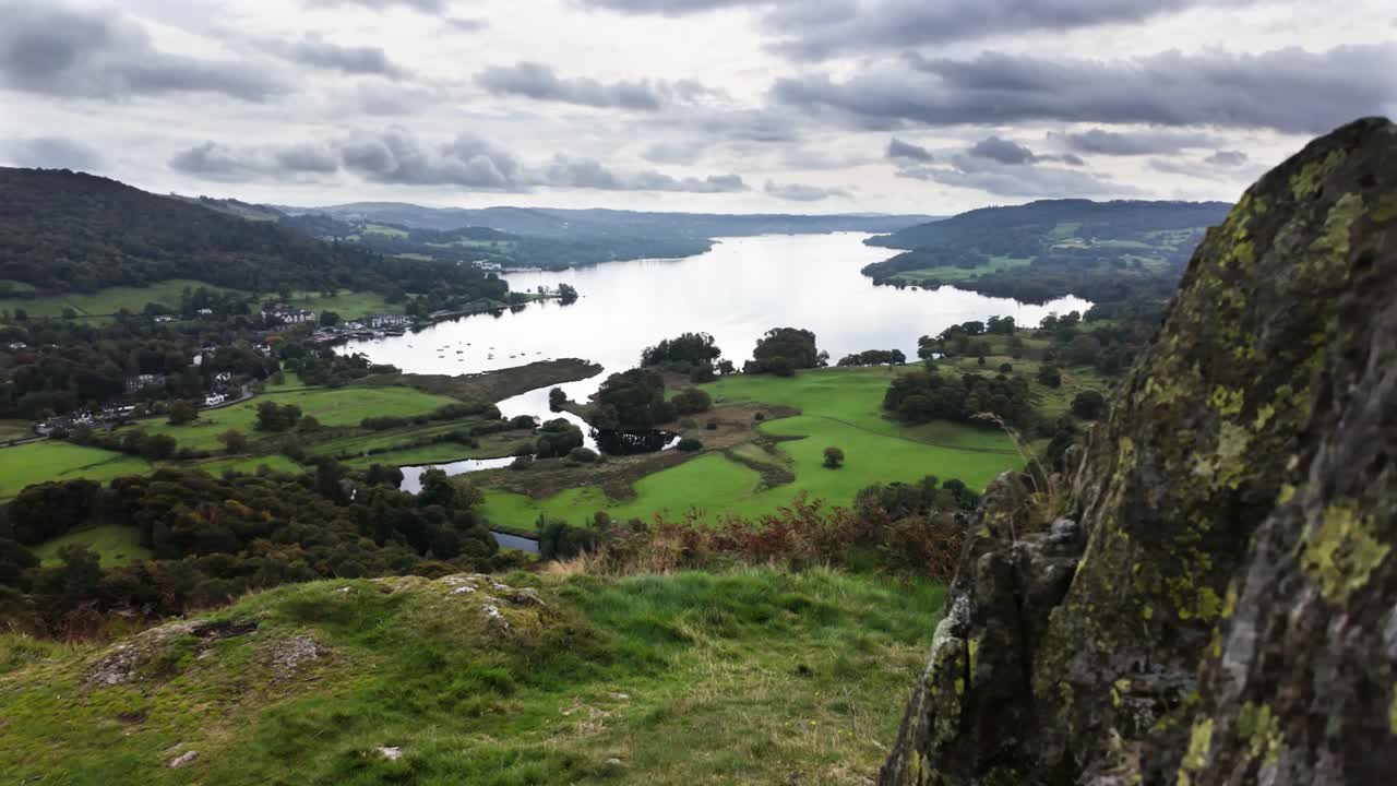 Moving past rocks on a hilltop to reveal Lake Windermere in the distance. Lake District, UK