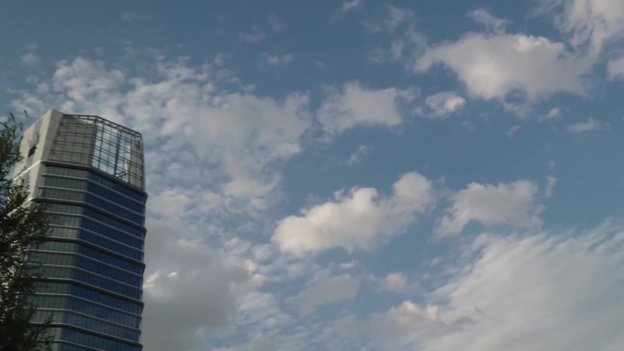Uma tomada time-lapse ampla de ângulo baixo captura nuvens dramáticas e fofas flutuando por um céu azul de verão acima de um moderno arranha-céu de escritórios de vidro no Infopark Budapeste, Hungria, em uma tarde ensolarada.