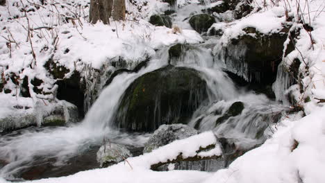 zoom out from a close up of a small waterfall flowing over rocks and ice, in a snow covered forest in alaska