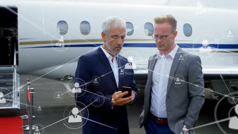 Network-of-profile-icons-against-two-caucasian-businessmen-using-smartphone-at-airport-runway