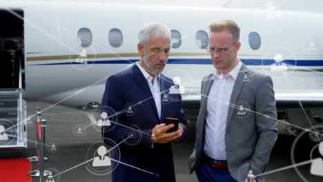 network of profile icons against two caucasian businessmen using smartphone at airport runway