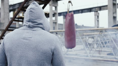 vista trasera de un hombre caucásico con ropa deportiva golpeando un saco de boxeo al aire libre en una fábrica abandonada en una mañana nublada