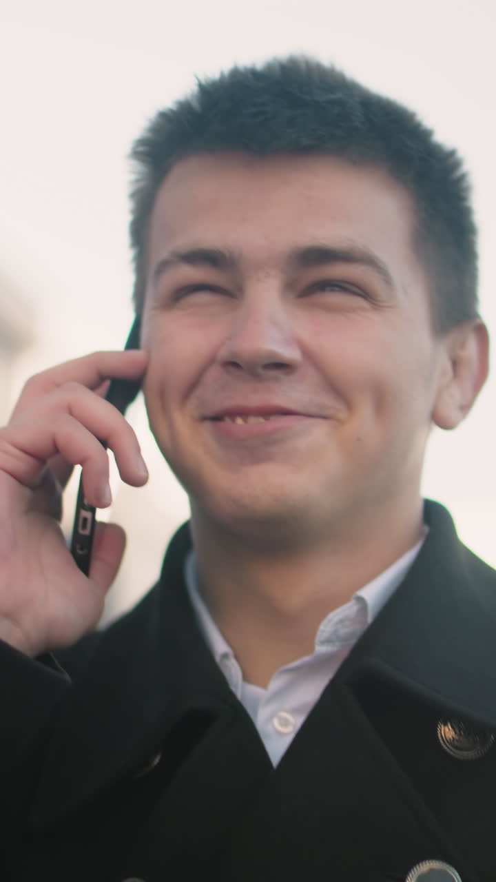 검은 코트를 입은 남자가 현대적인 유리 건물, 부드러운 빛, 그리고 즐거운 분위기를 가진 활기찬 도시 지역에서 손짓을 하면서 흥분하여 전화로 행복하게 이야기하고 있습니다.