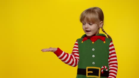 Kid-girl-in-Christmas-elf-Santa-helper-costume-points-fingers-at-blank-space-shows-advertising-area