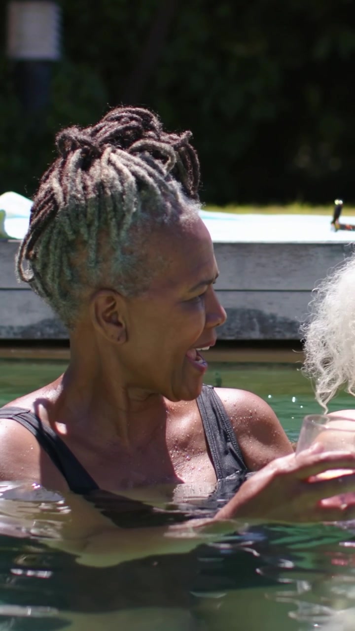 Video verticale: Anziana con top da bagno nero che porta bicchieri senza stelo di rosé e brinda con un'amica in piscina.