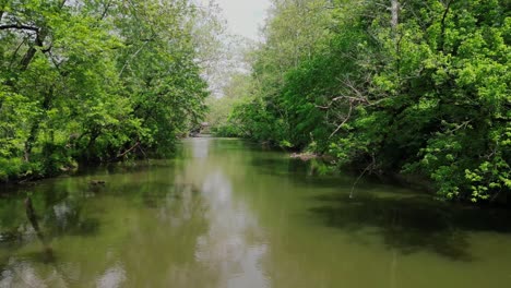 Drone-flying-over-river-water-with-green-trees-on-bank-in-Indiana