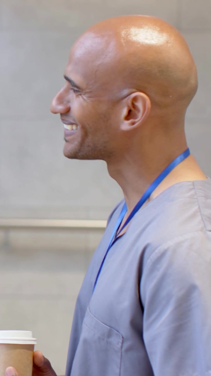 Vertical video: Smiling male in scrubs, lanyard in hospital reacting to offscreen voice, taking cup
