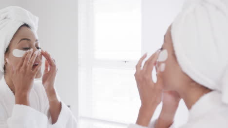 Biracial-woman-applying-under-eye-patches-looking-in-mirror-in-bathroom,-slow-motion