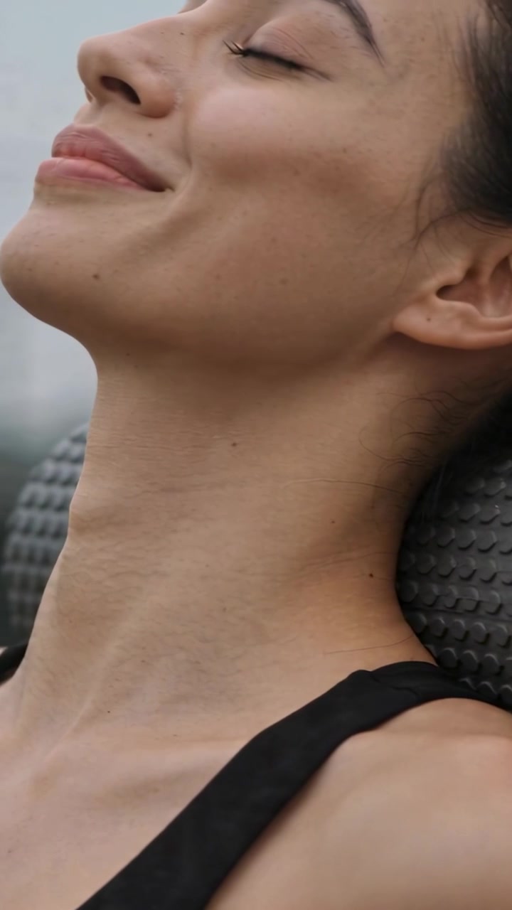 Video verticale: Donna con canotta nera che si adagia, reclinando la testa all'indietro e sorridendo su un cuscino cervicale, mentre si rilassa sul tetto