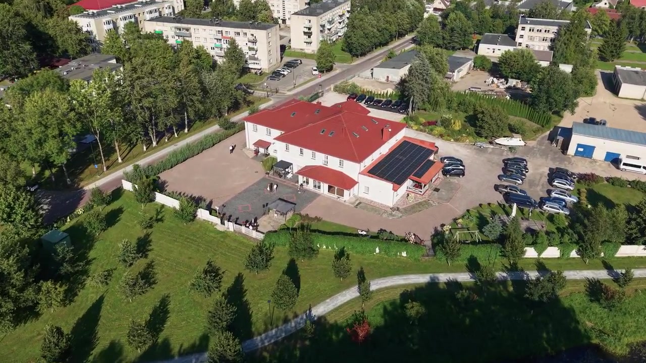 Vídeo suave de drone revela uma área suburbana tranquila com edifícios de telhados vermelhos, ruas arborizadas, carros estacionados e vegetação exuberante sob a luz clara do sol da tarde. Imagens aéreas amplas.