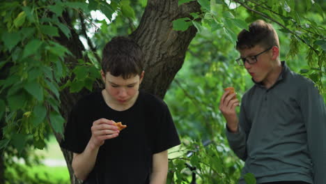 siblings enjoy a break while eating snacks under a tree in a peaceful forest, one of the boys leans on the tree while the other stands, both surrounded by lush greenery