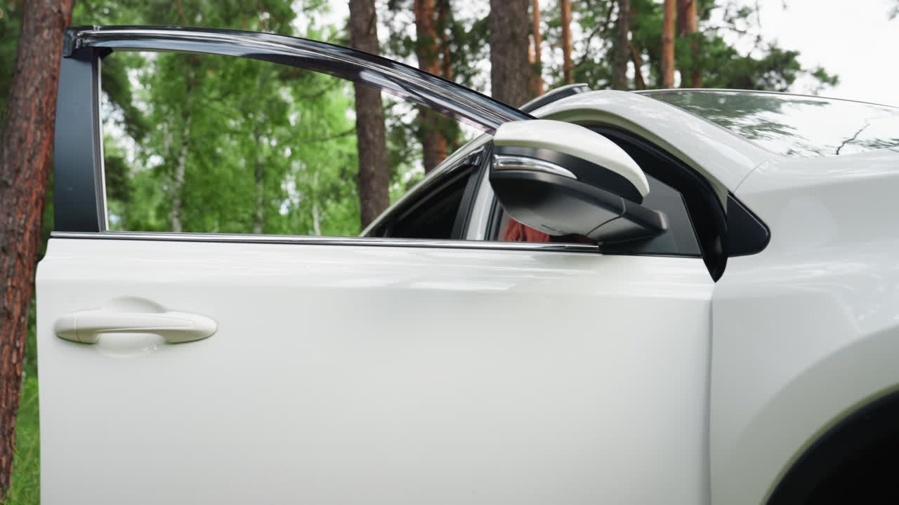 Lower view of trendy woman with long red hair smiling as she opens car door, sits inside and closes it, parked in lush green arboretum surrounded by tall trees, calm atmosphere with natural daylight
