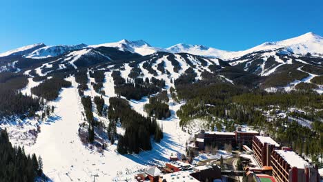 aerial drone view of people skiing and riding chairlifts down ski slope next to resort hotels and pine tree forest with snow