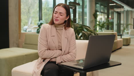 mujer de negocios caucásica decepcionada usando una computadora portátil y sintiendo un dolor de estómago repentino en el vestíbulo de la oficina