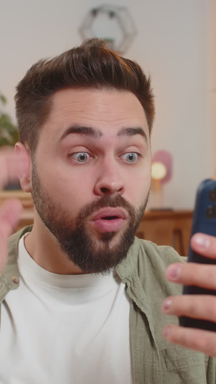 Surprised caucasian brunette man using smartphone winning lottery sitting on sofa couch at home