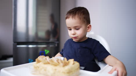 lovely baby sitting at high chair in the kitchen. mother puts the box with a cake in front of her son.