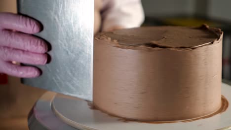 pastry chef woman is smearing chocolate cream on sponge cake with metal spatula on rotating cake stand in bakehouse.