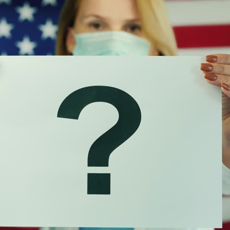 a woman in a medical mask holds a poster with a question mark against the background of the american flag 1