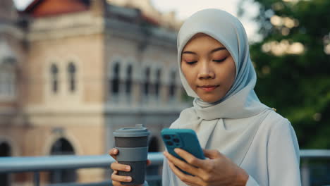 young muslim woman using smartphone outdoors with coffee cup