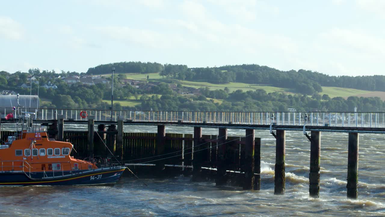 An orange lifeboat approaches a pier in rough waters near the Scottish Highlands coastline