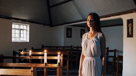young woman in a white dress stands with her eyes closed in a dimly lit church, three candles burning on a wooden pew in the foreground, creating a serene atmosphere of prayer and reflection
