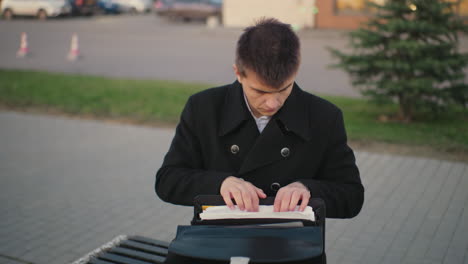 businessman seated outdoors after work, going through documents in urban setting, office building in background, street light and parked car at twilight