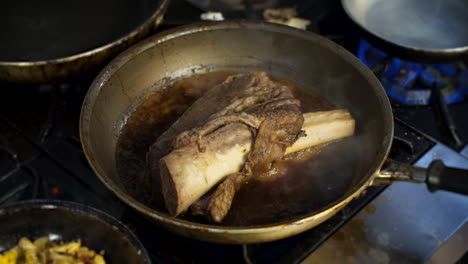 Skillet-simmering-beef-rib-on-stove-in-commercial-restaurant-kitchen,-beer-braising-beef-in-skillet,-slow-motion-HD