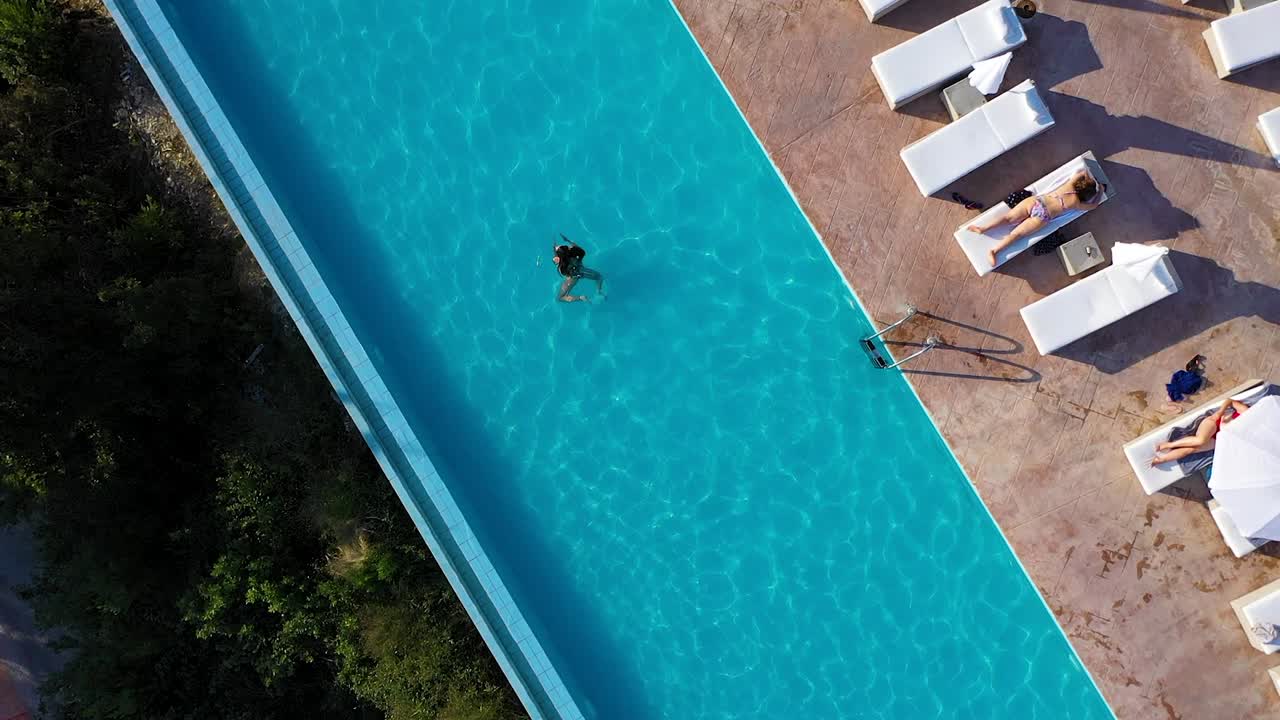 Aerial view young woman swims through a hotel pool, environment is seen also.