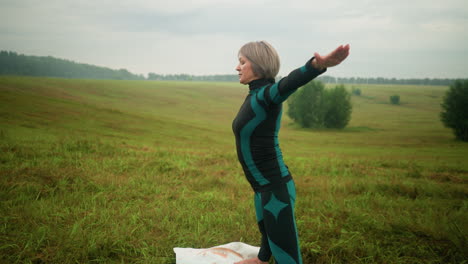 side view of woman in green and black suit standing on yoga mat, practicing yoga triangle pose with arms extended and legs apart, set against a serene grassy landscape with trees and greenery