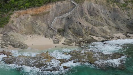 Aerial-pan-shot-of-a-stairs-leading-to-ocean-with-waves-hitting-the-coast-of-Baie-Rouge-Beach-at-Saint-Martin