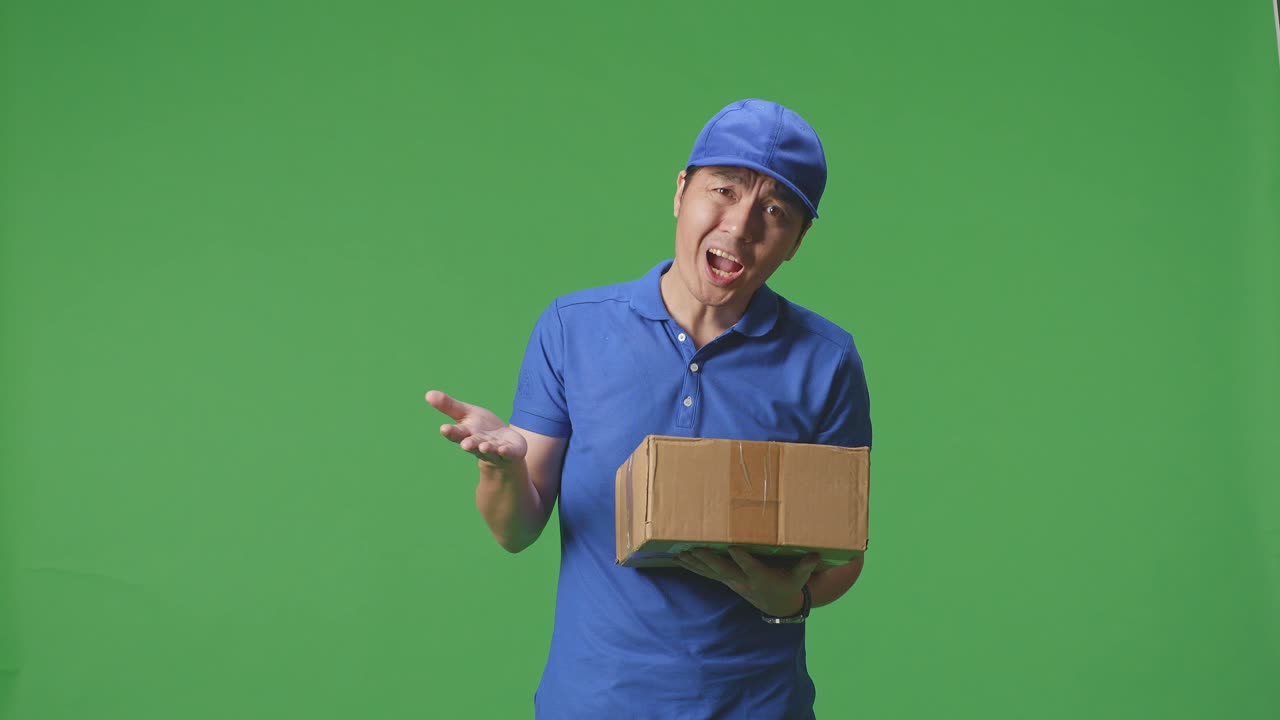 Asian Male Courier In Blue Uniform Showing Puzzled Gesture And Saying What While Delivering A Carton On Green Screen Background In The Studio