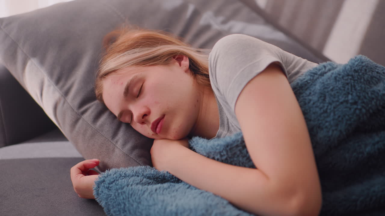 Sleepy young woman nestled under thick textured blanket gently turns right while lying on couch, morning light casting calm shadows across soft surface in quiet relaxed indoor atmosphere
