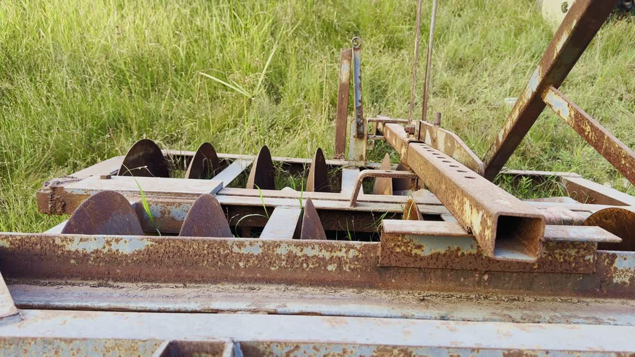 A static shot capturing an old rusty disc harrow lying idle in tall green grass, its metal surfaces covered in rust and dust — a quiet reminder of once-active agricultural machinery now aging